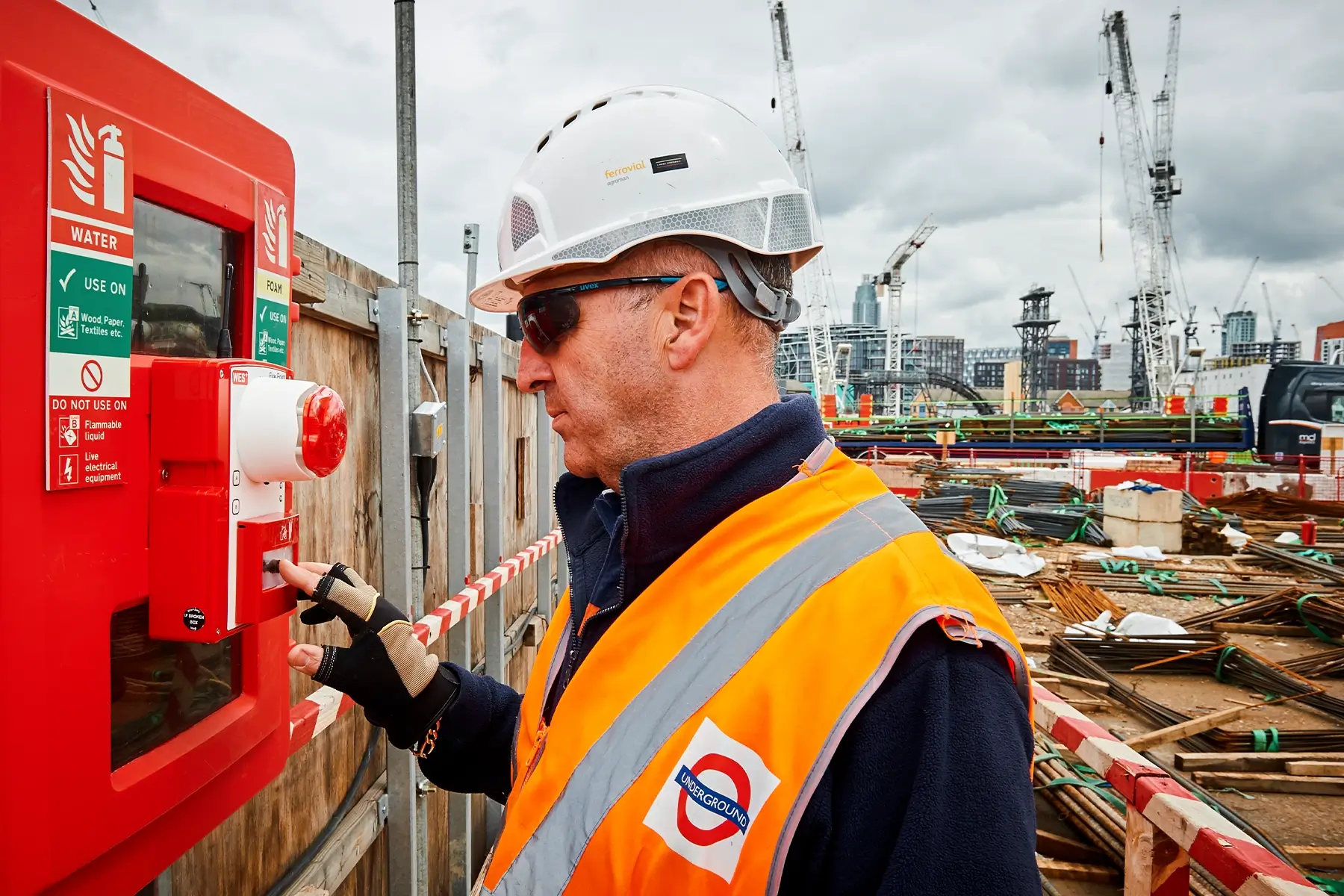 Construction worker pressing a manual call point on a red WES+ wireless fire alarm unit for the Northern Line Extension Fire Safety.