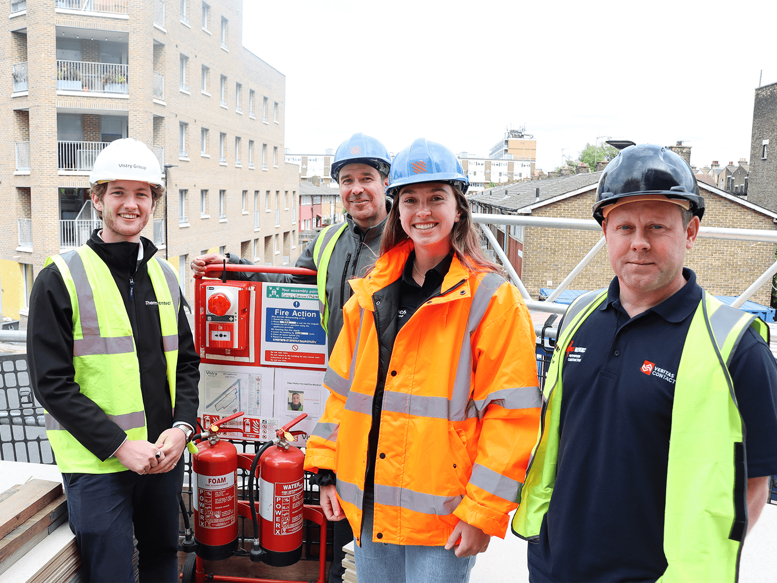 Quatre ouvriers du bâtiment en tenue de sécurité autour d'un poste d'incendie mobile rouge équipé d'extincteurs et d'une alarme incendie.