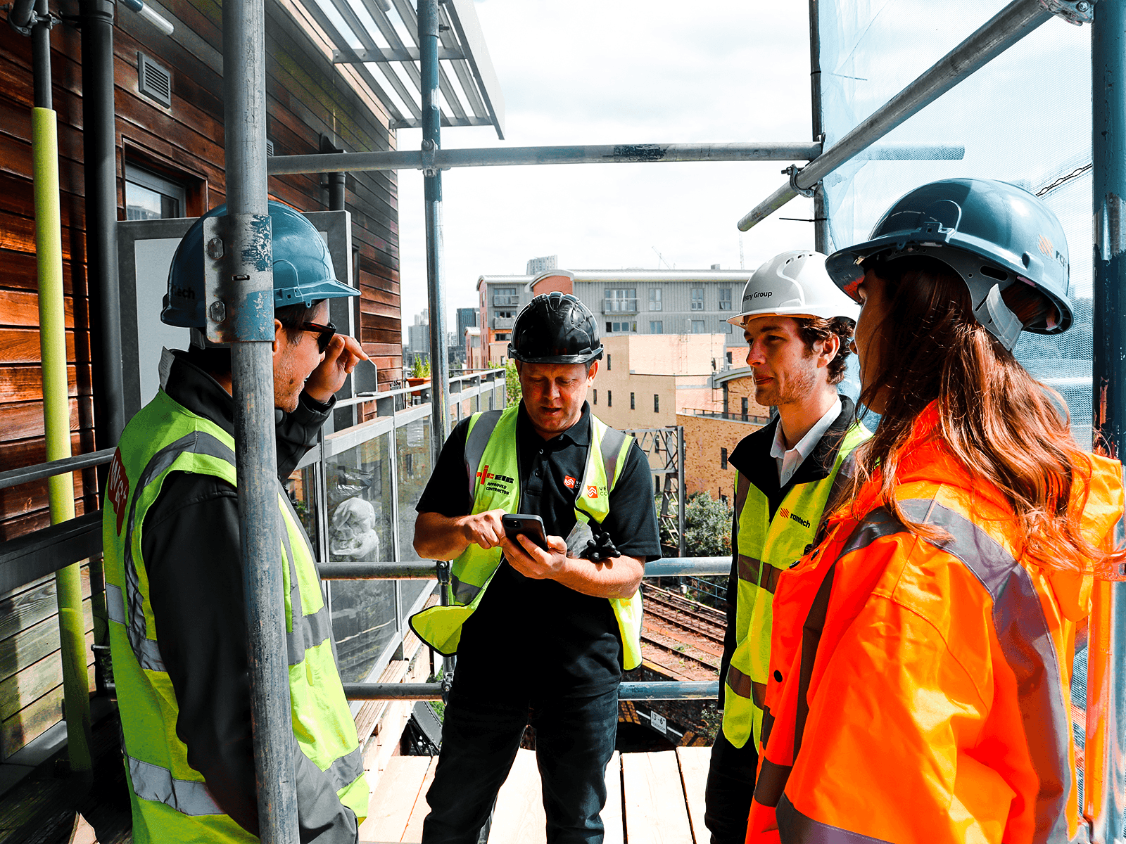 Quatre professionnels de la construction en tenue de haute visibilité se sont rassemblés sur le balcon d'un échafaudage, regardant un smartphone tenu par un homme.