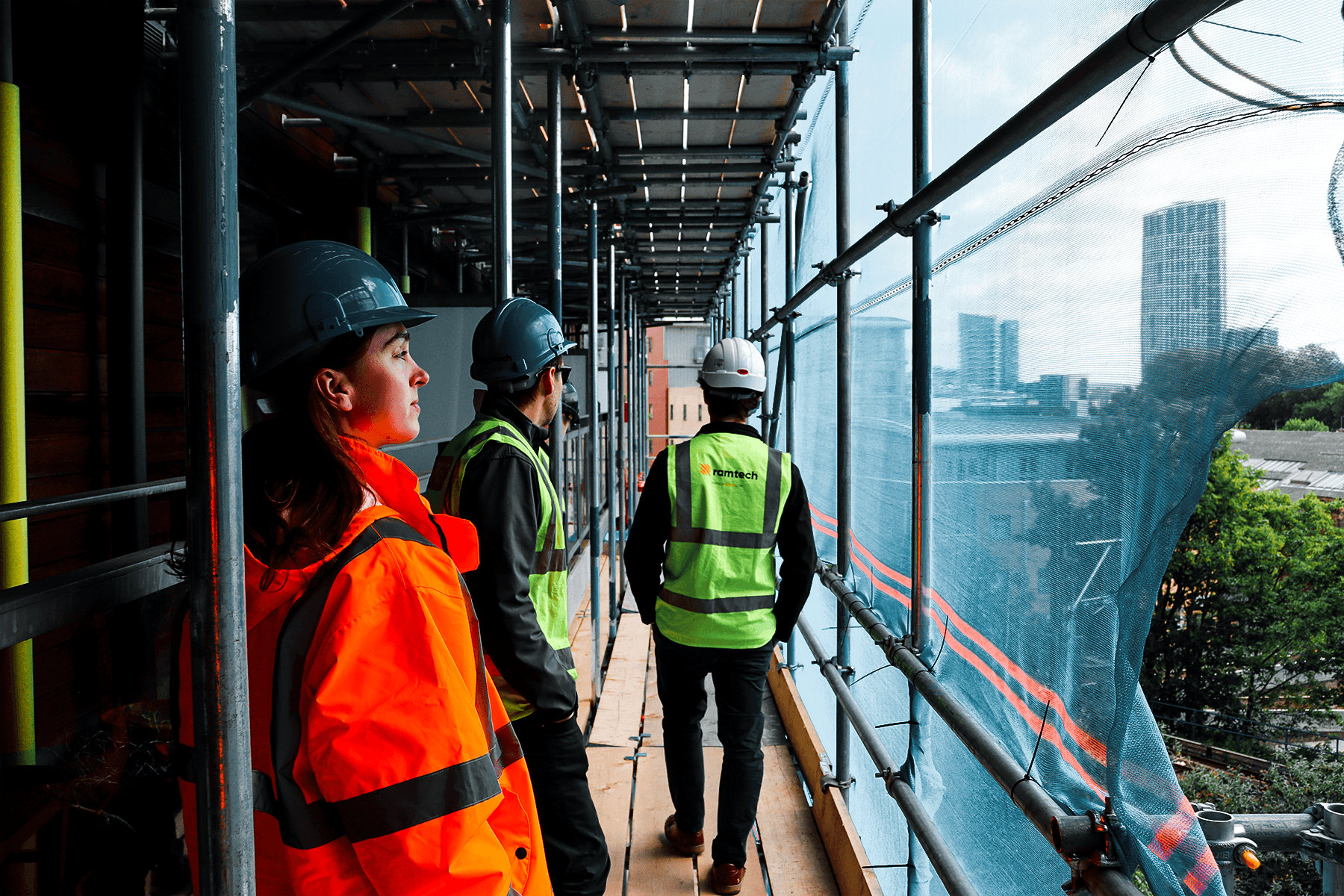 Construction workers standing on a high-level scaffold walkway overlooking city skyscrapers in the distance.