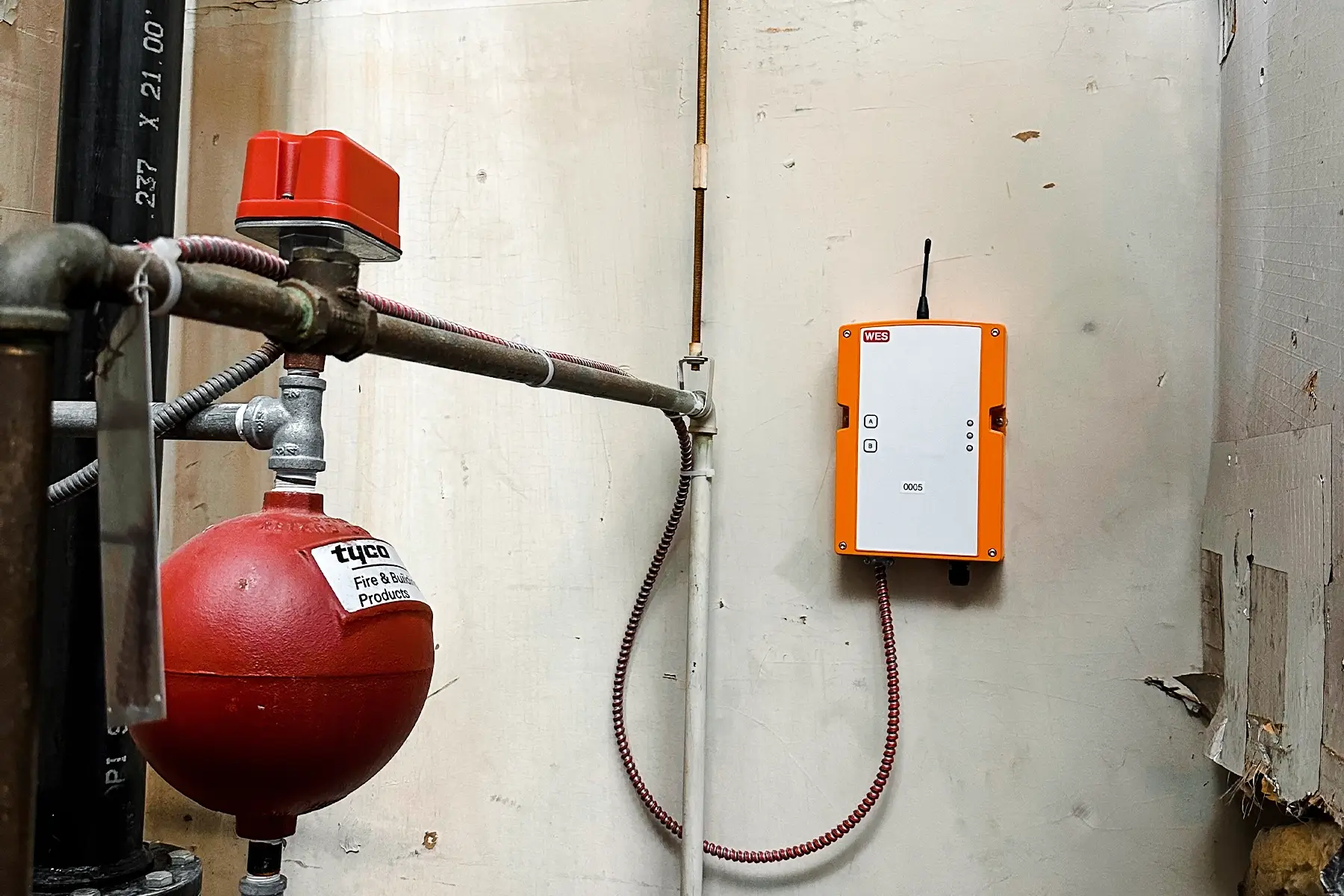 Two men installing or inspecting a WES3 control panel (orange box) next to a large red fire alarm panel. The WES3 is a Wireless temporary fire alarm system.