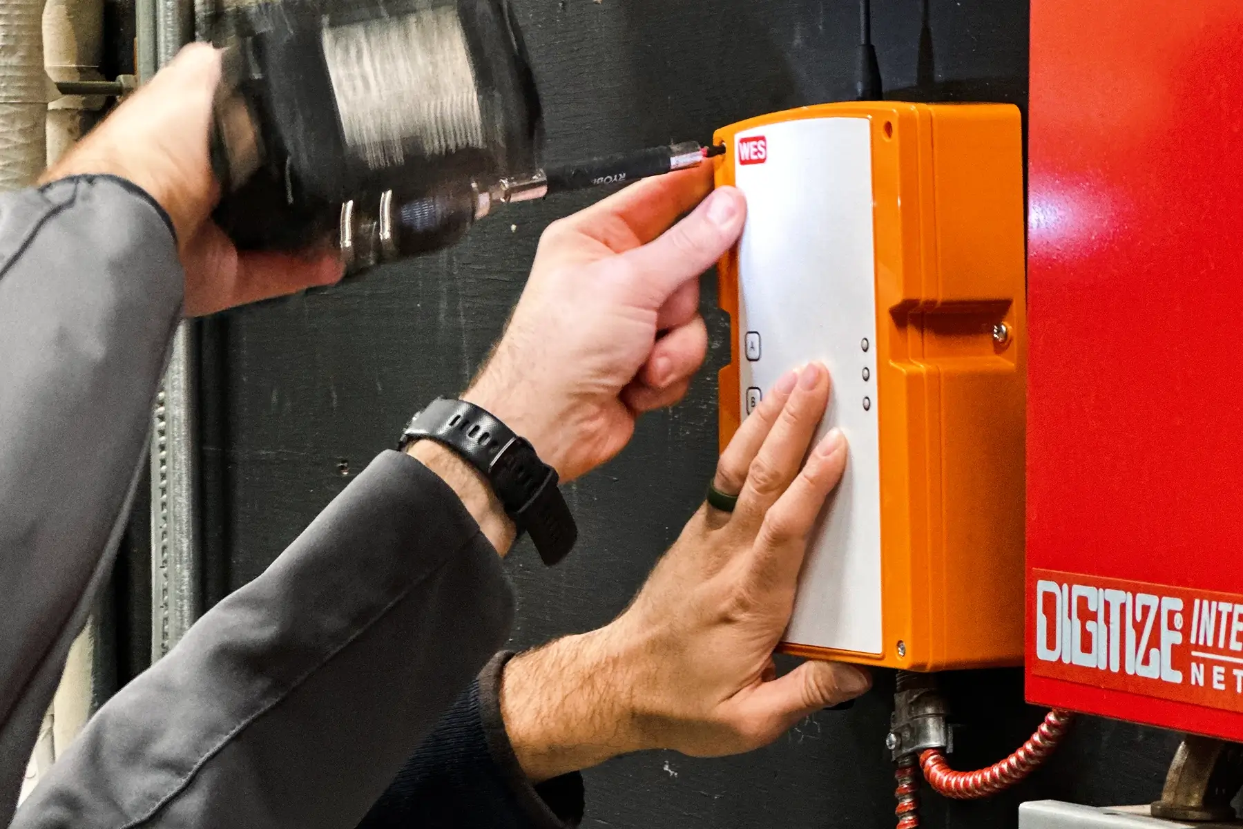 A close-up of a technician connecting an external component to an orange WES3 unit. The system is a Wireless temporary fire alarm system.