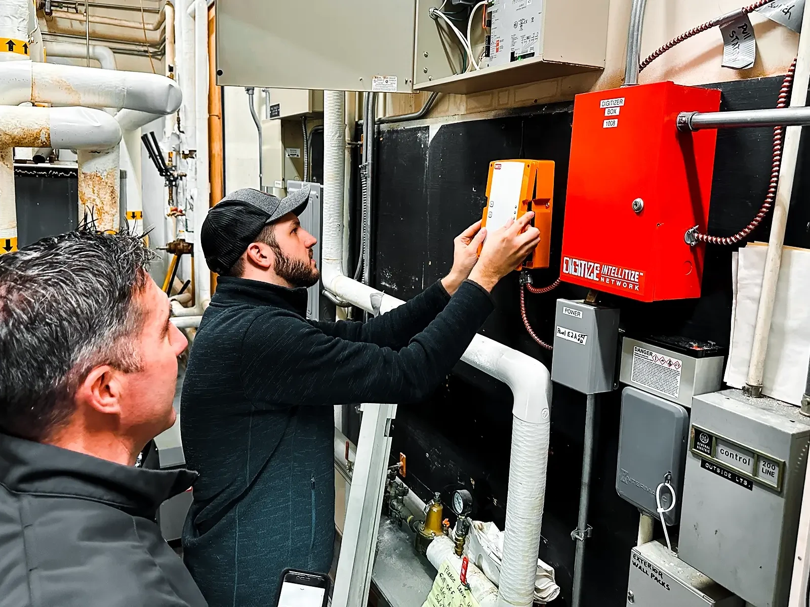 Un homme portant une casquette de baseball et une barbe se concentre sur le panneau de contrôle orange WES3 monté sur un mur sombre. Il s'agit d'un composant du système d'alarme incendie temporaire sans fil.