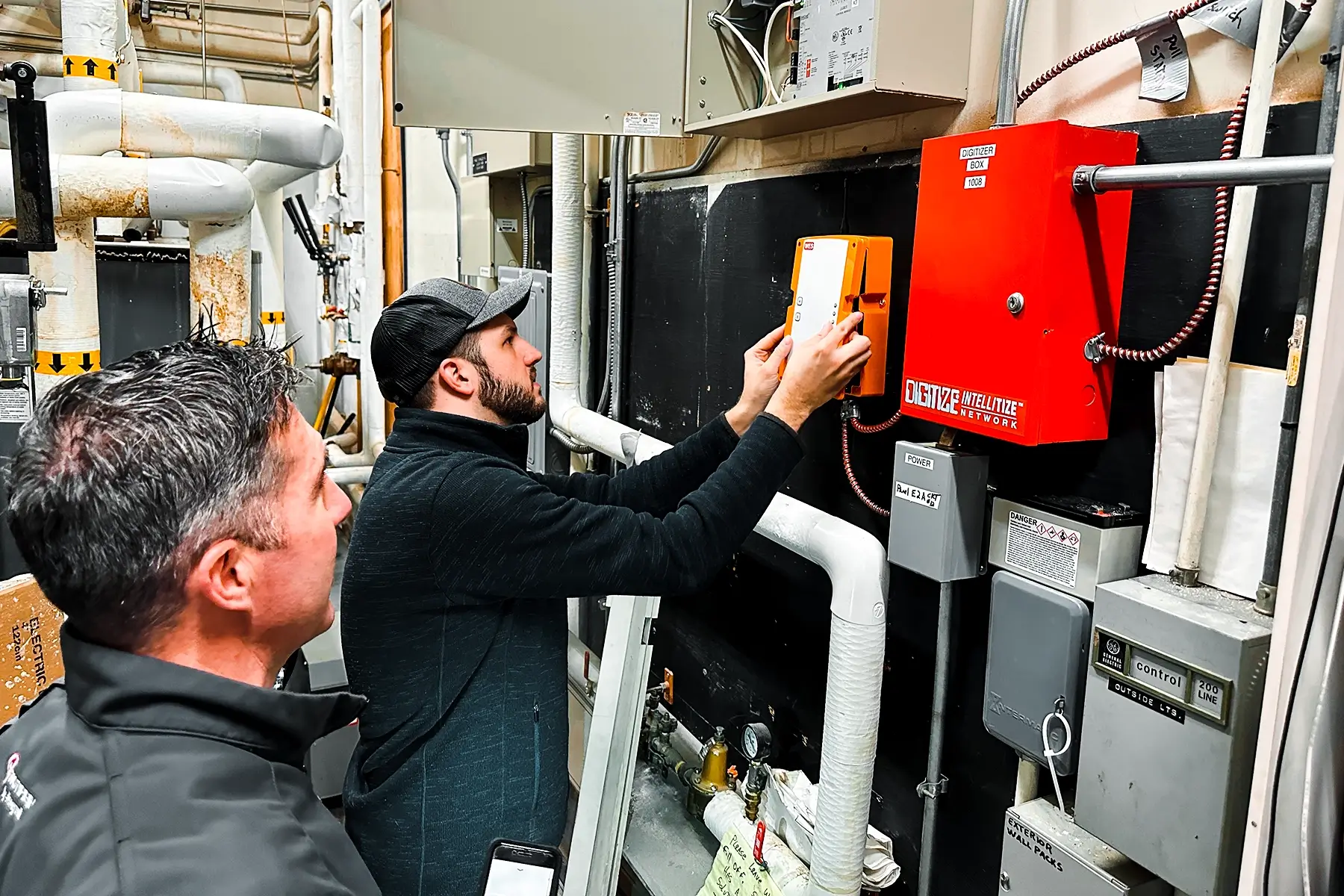 A man wearing a baseball cap and a beard focusing on the orange WES3 control panel mounted on a dark wall. This is a component of the Wireless temporary fire alarm system.